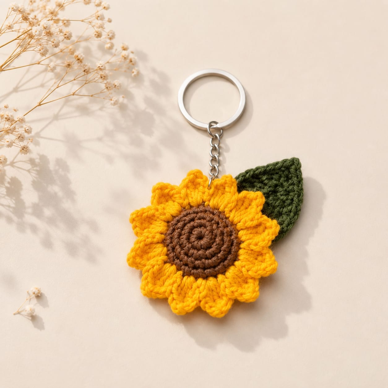 A crocheted sunflower keychain with a silver metal split ring and chain, featuring a yellow and brown flower body and a green leaf, displayed on a beige background with dried flowers.