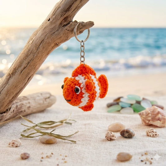 A crocheted orange and white fish keychain hanging from a driftwood branch on a sandy beach with shells and the ocean in the background.