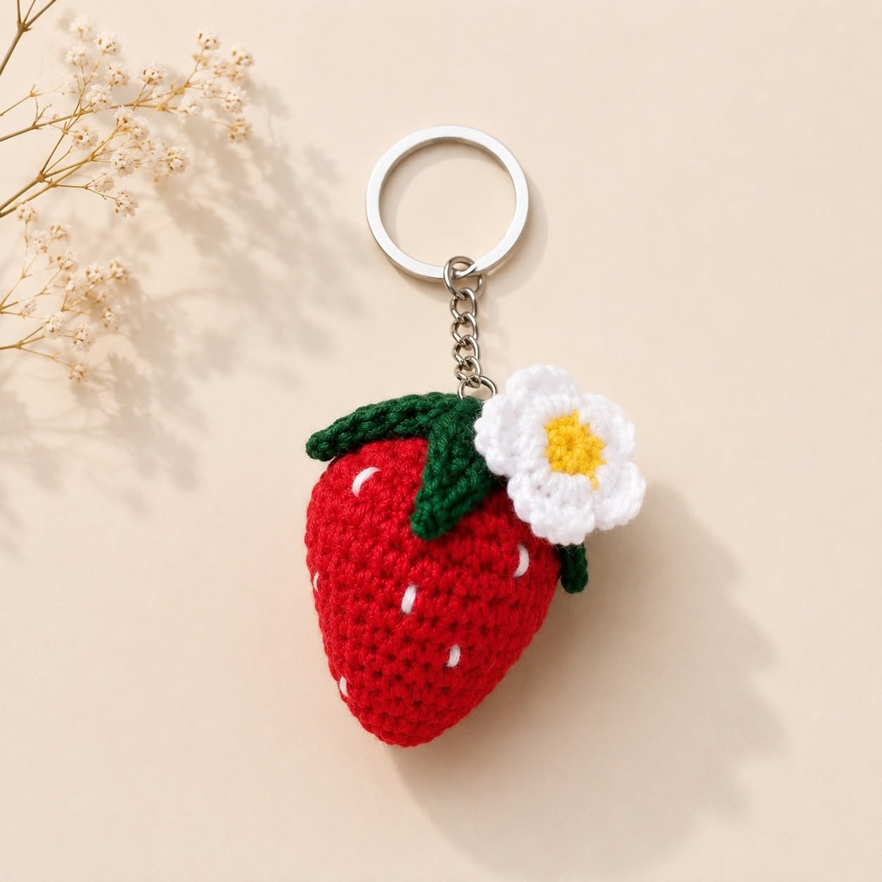 A crocheted strawberry keychain with a silver metal split ring and chain, featuring a red body, green leafy top, and a white flower with a yellow center, displayed on a beige background with dried flowers.